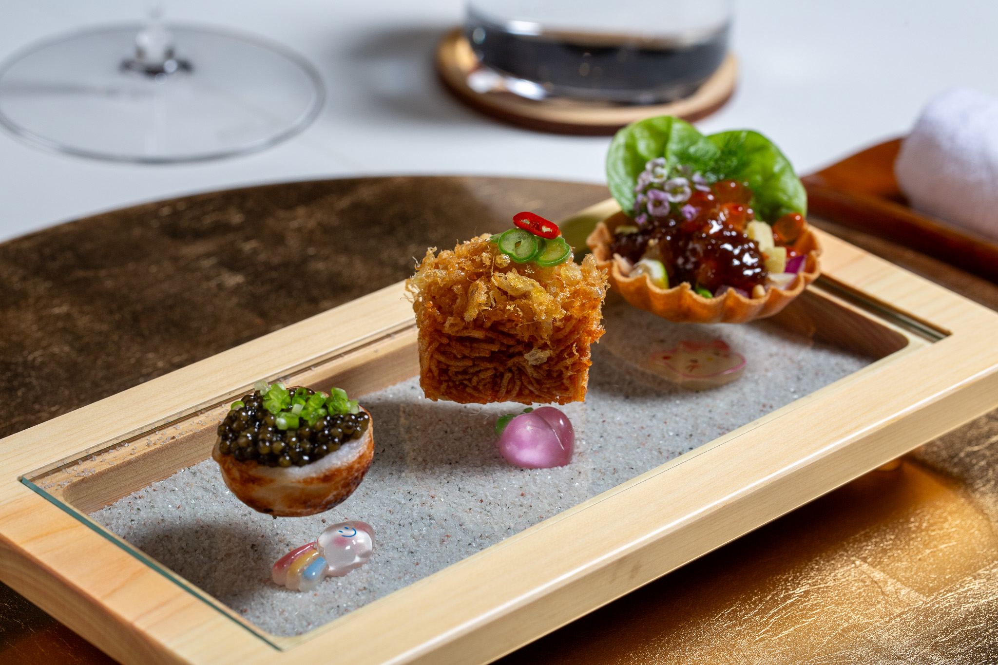Three small desserts on a wooden tray with a mirror surface, garnished with colorful toppings and sauces.