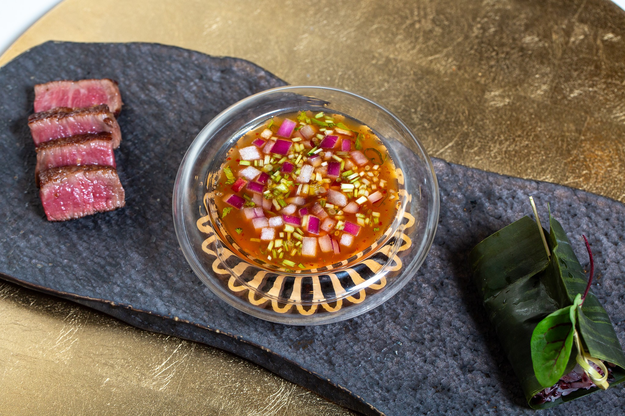 Slices of cooked meat, a bowl of dipping sauce with chopped vegetables, and a wrapped leaf on a dark surface.