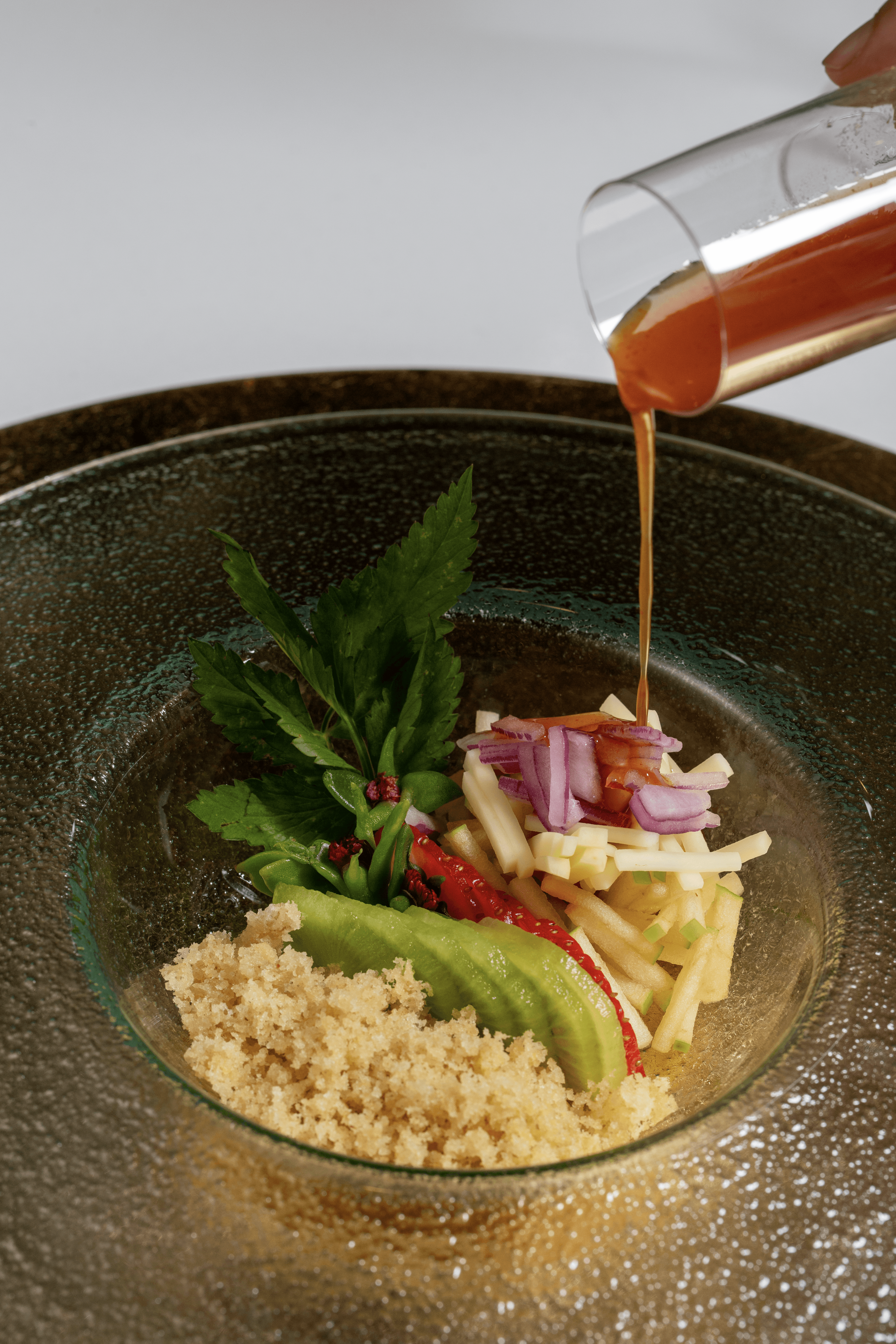 Liquid is poured from a glass into a bowl with vegetables and grains, on a dark plate.
