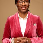 Woman in pink velvet suit with glasses and pearl bracelet, smiling, standing against a beige background.