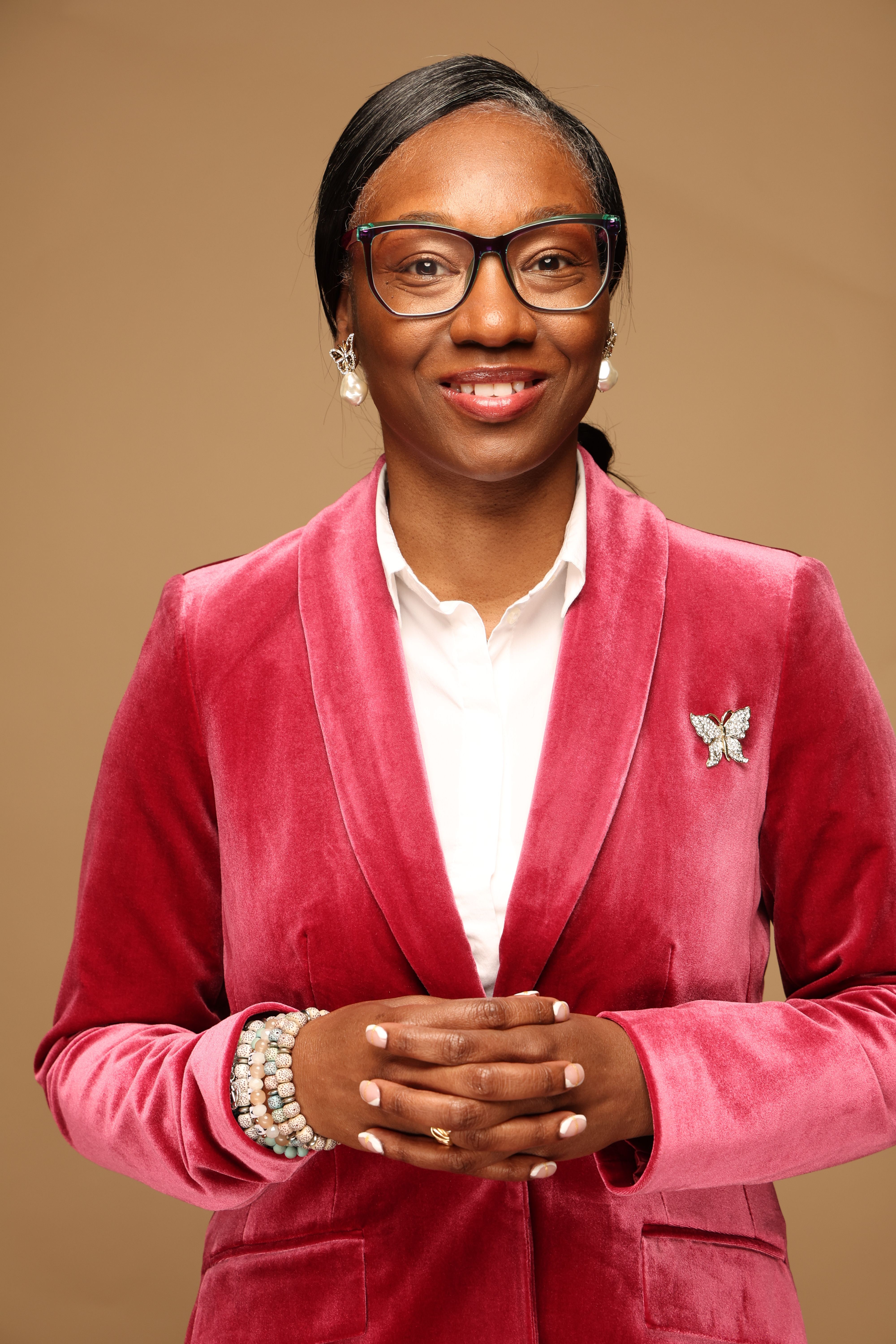 Woman in pink velvet suit with glasses and pearl bracelet, smiling, standing against a beige background.