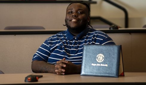 Young person sitting at a desk with a laptop and a blue folder, smiling, in a classroom or lecture hall.