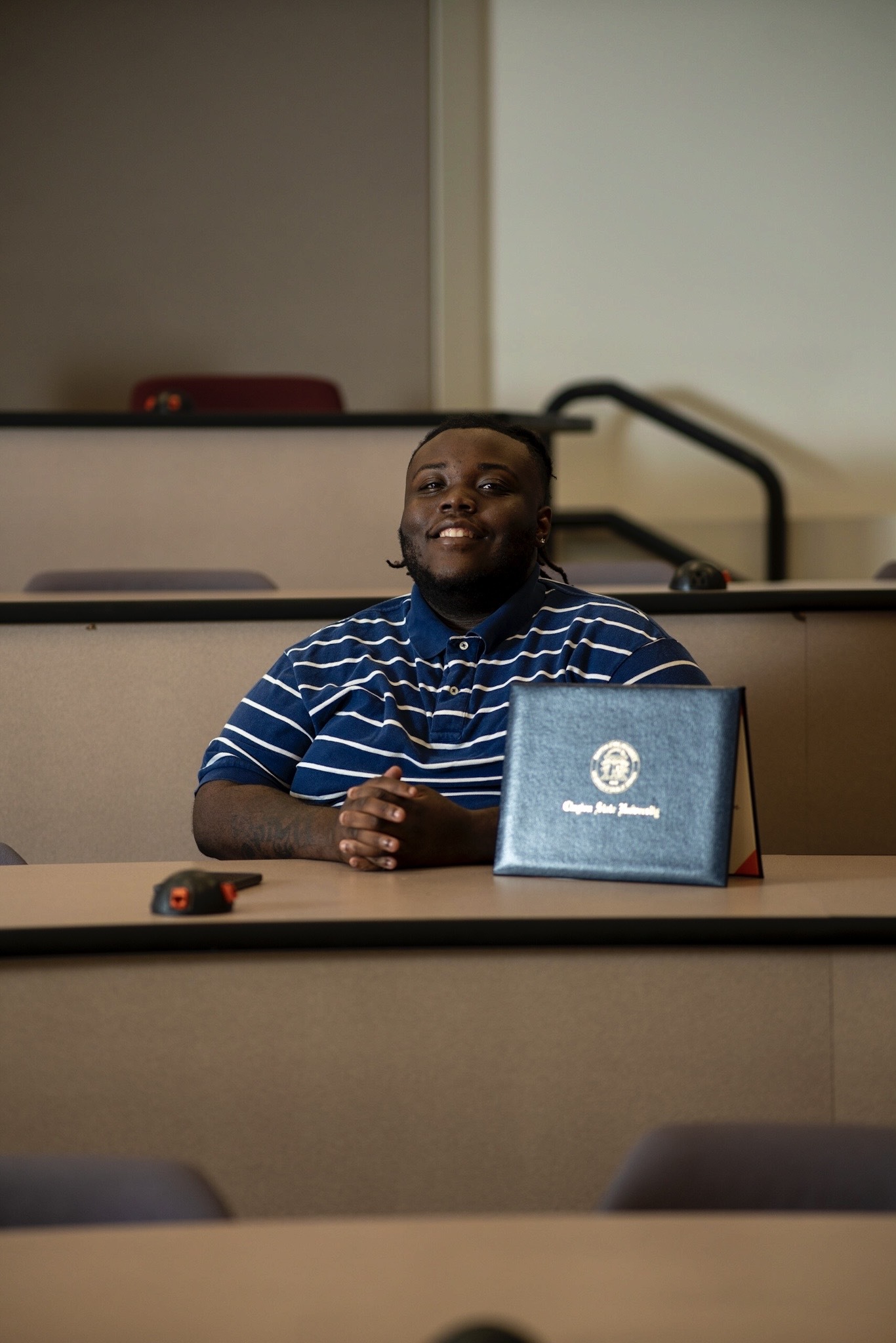 Young person sitting at a desk with a laptop and a blue folder, smiling, in a classroom or lecture hall.