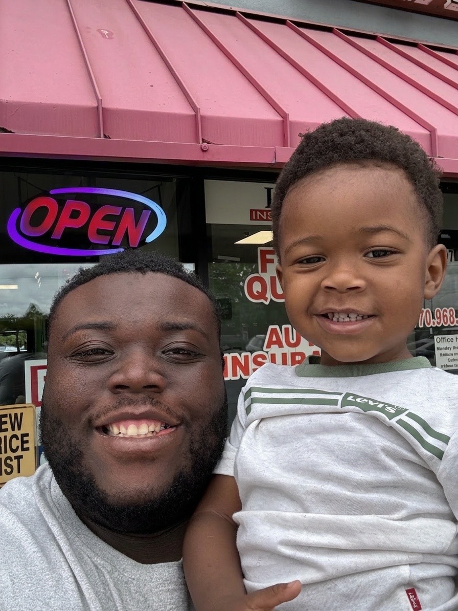 Two smiling males, one adult and one child, standing outside a store with an open sign in the window.
