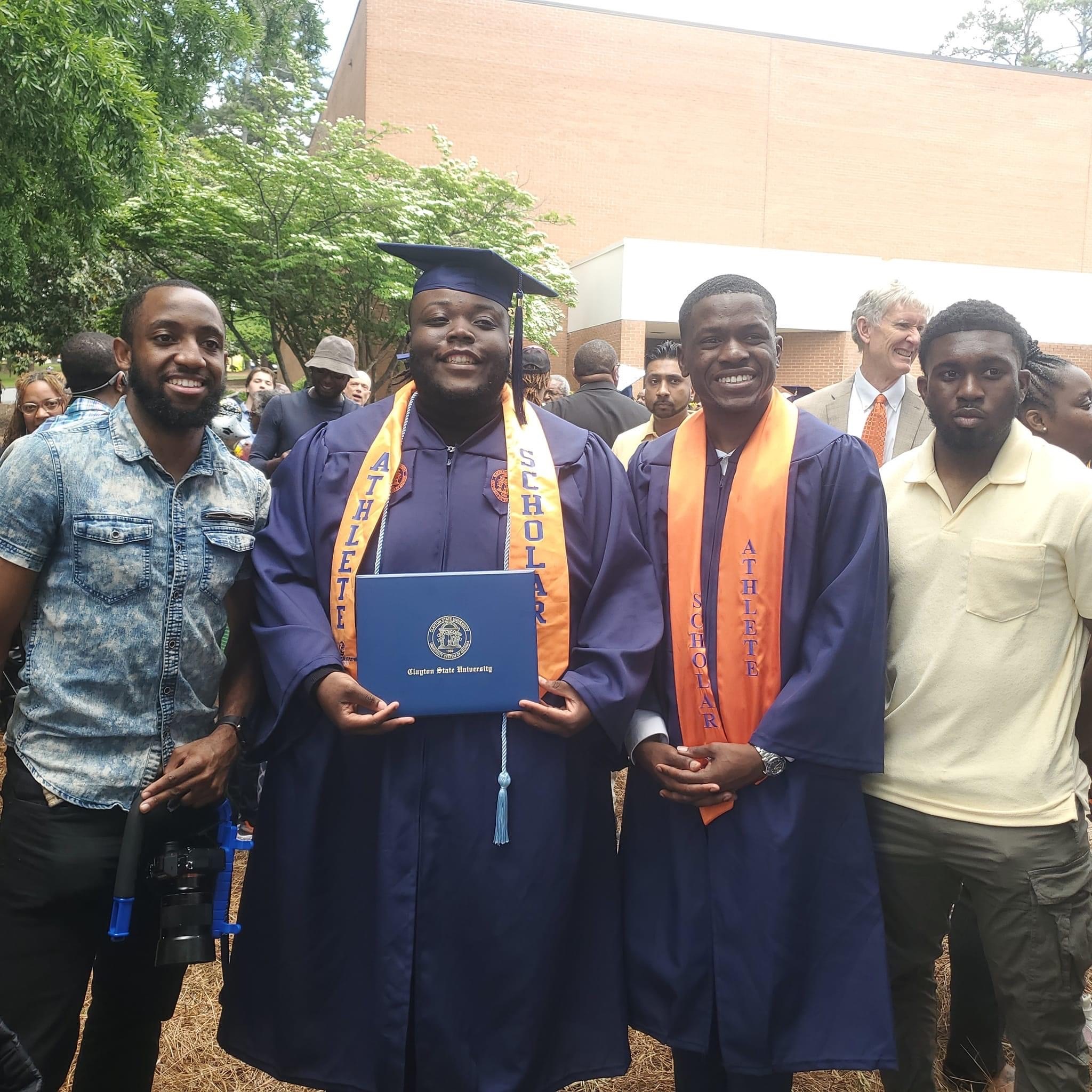 Group of people celebrating graduation outdoors, with two graduates in caps and gowns holding a diploma, smiling, surrounded by friends and family.