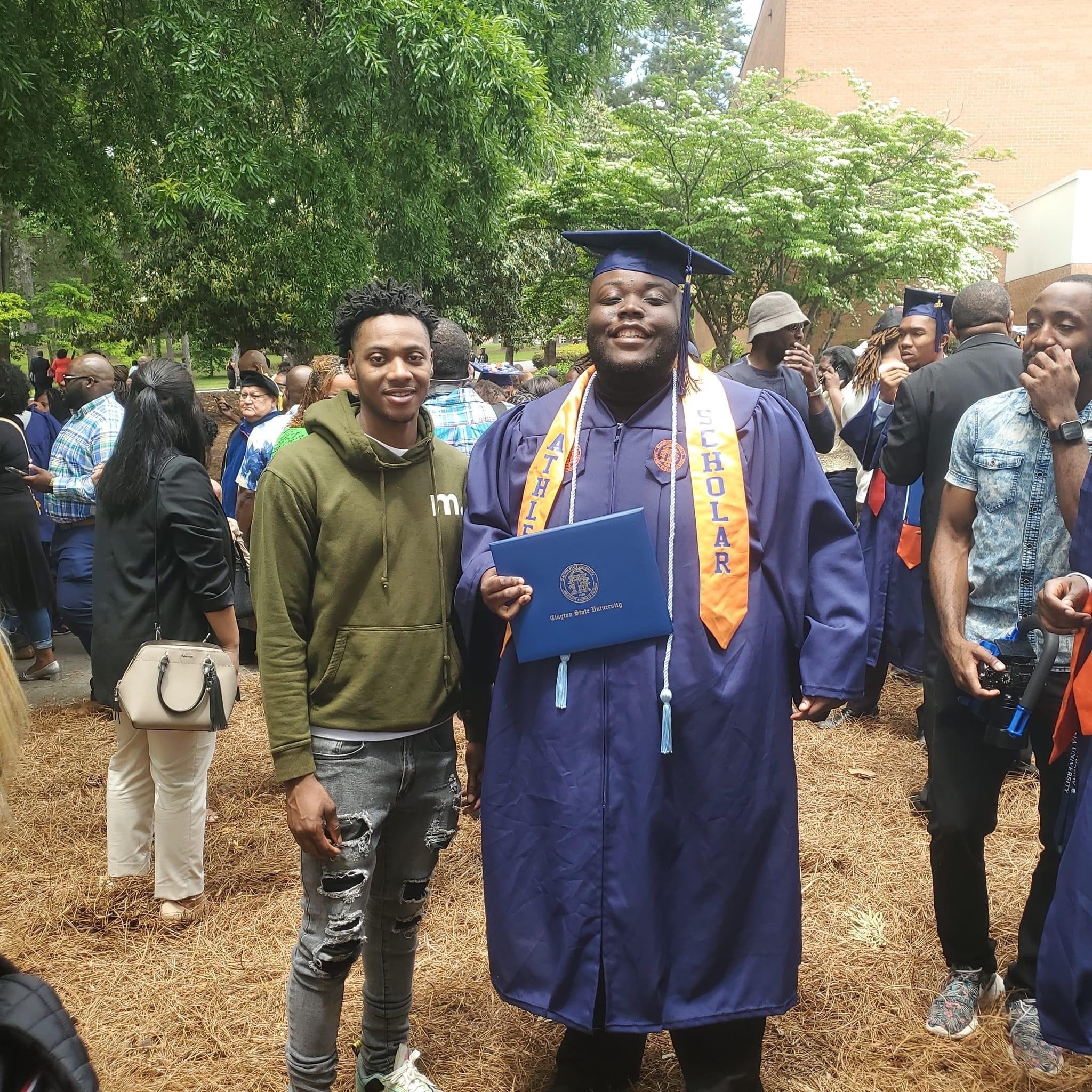 Graduate in cap and gown holding diploma, smiling, surrounded by people outdoors, trees in background.