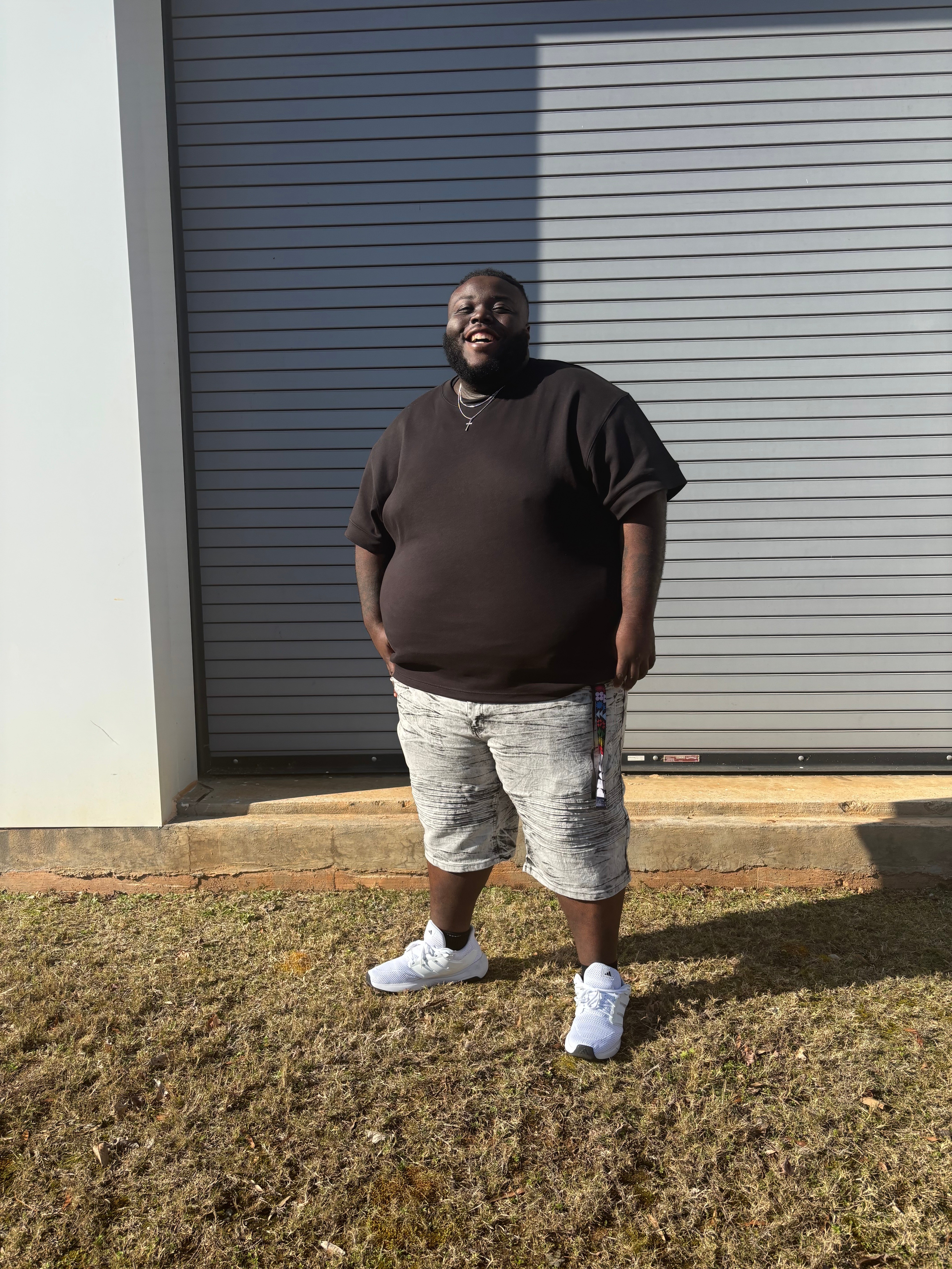 Person standing outdoors in front of a gray garage door, smiling, wearing a black shirt, patterned shorts, and white sneakers.
