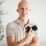 Smiling man holding a camera, arms crossed, in a light-colored shirt, standing indoors with a blurred background.