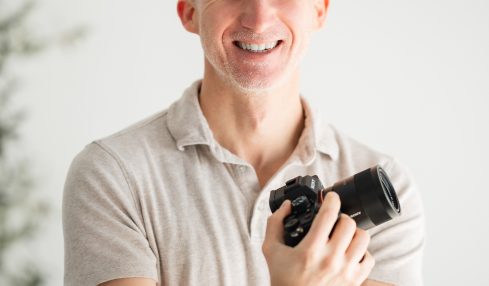 Smiling man holding a camera, arms crossed, in a light-colored shirt, standing indoors with a blurred background.