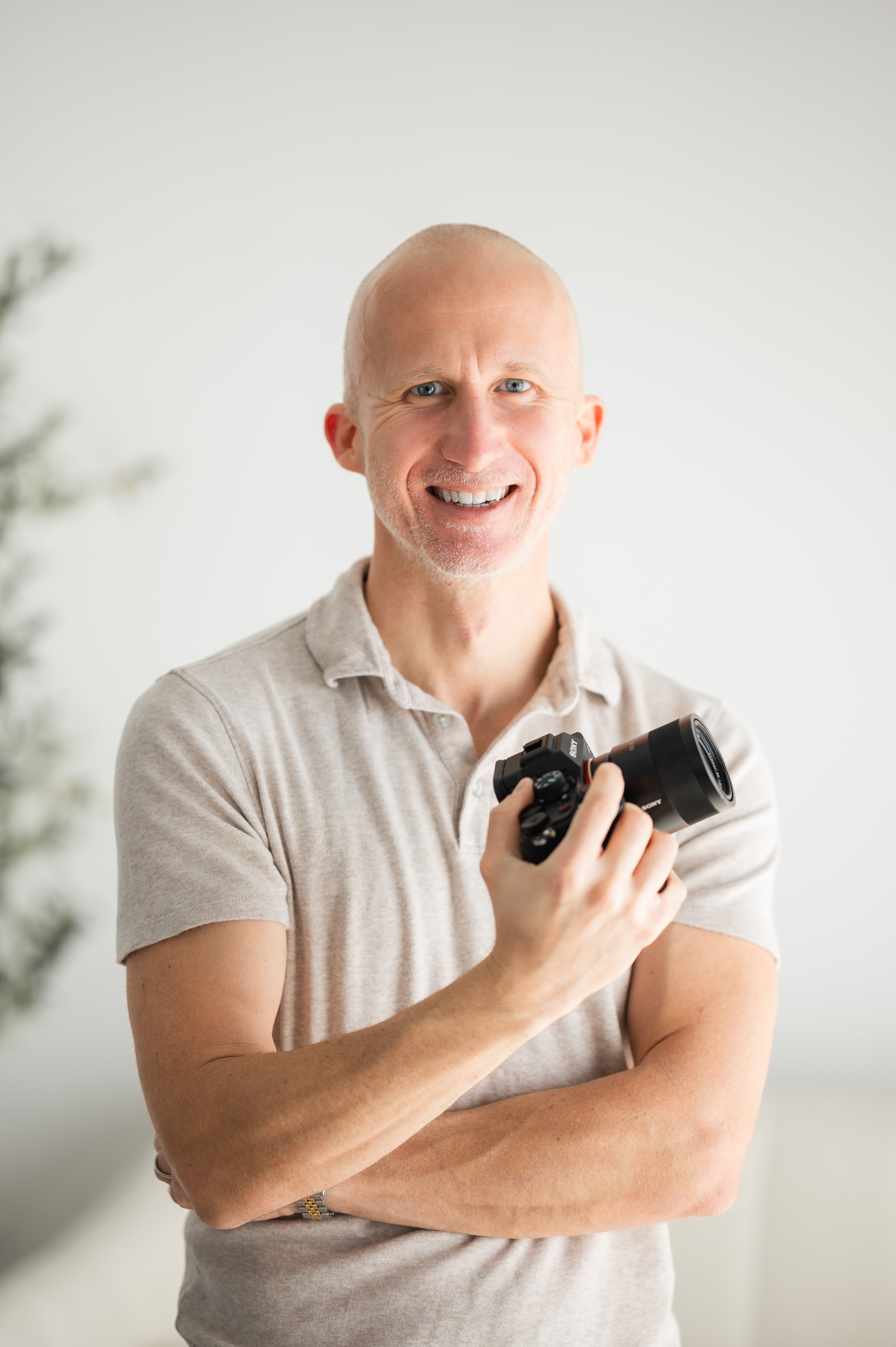 Smiling man holding a camera, arms crossed, in a light-colored shirt, standing indoors with a blurred background.