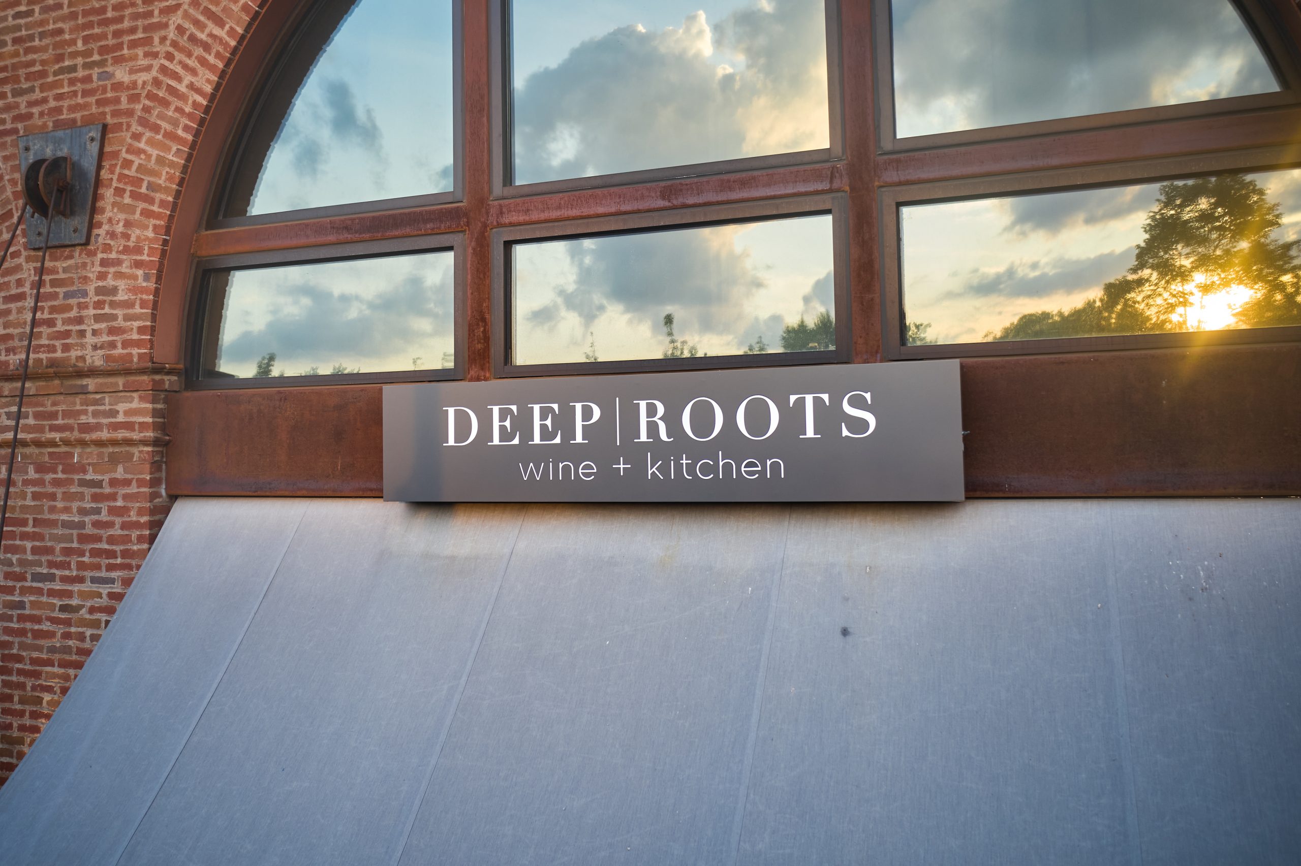 Sign for Deep Roots wine and kitchen restaurant outside a brick building with large arched windows, sunlight shining through clouds.