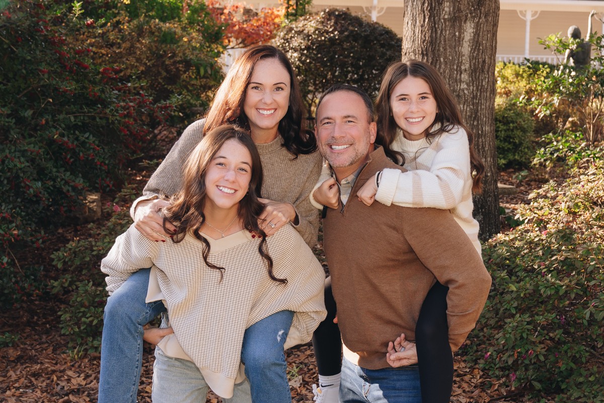 Four people smiling outdoors, two women, one man, and a girl, standing among trees and bushes with sunlight.