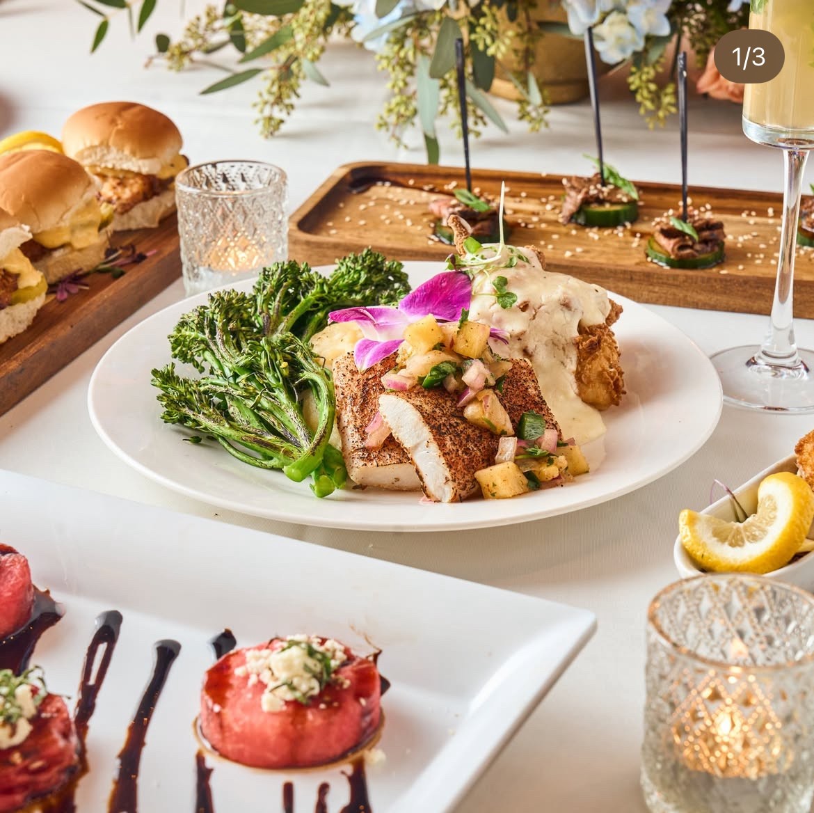 Plate with breaded meat, broccoli, and creamy sauce garnished with edible flowers, on a white table with drinks and food in background.