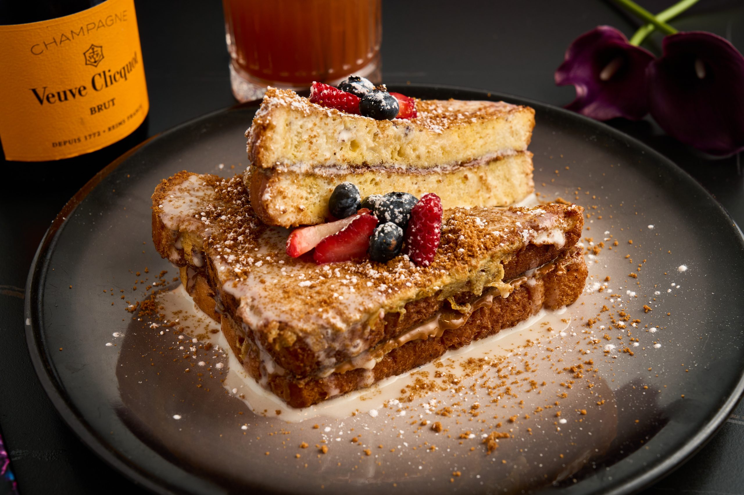 Two slices of French toast with berries and powdered sugar on a black plate, with a drink and purple flowers nearby.