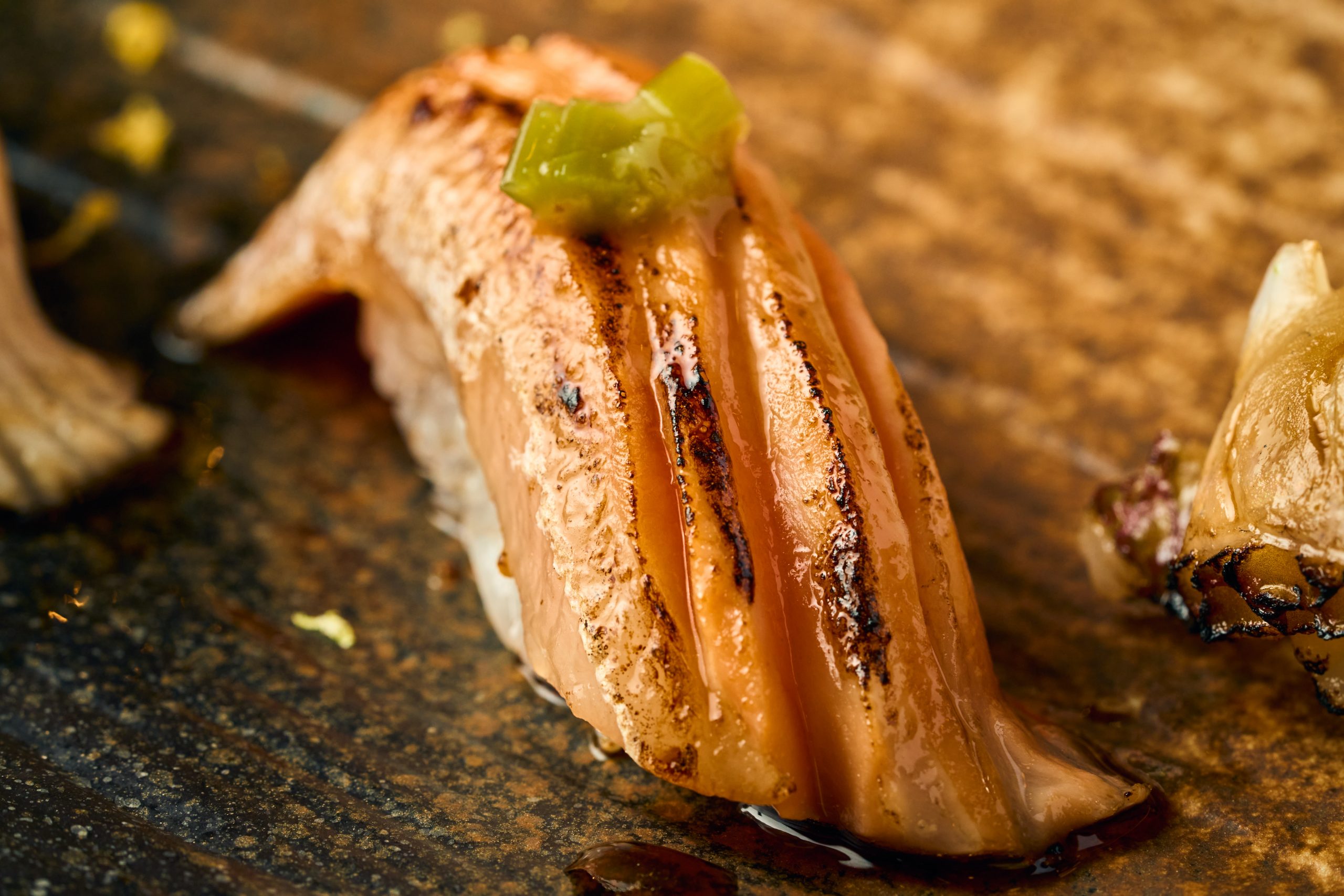 Close-up of a piece of sushi with fish, topped with green wasabi, on a wooden surface.