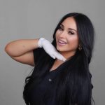 Woman with long black hair smiling, wearing a black top and white glove, posing with her hand near her face.