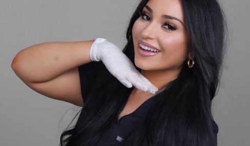 Woman with long black hair smiling, wearing a black top and white glove, posing with her hand near her face.