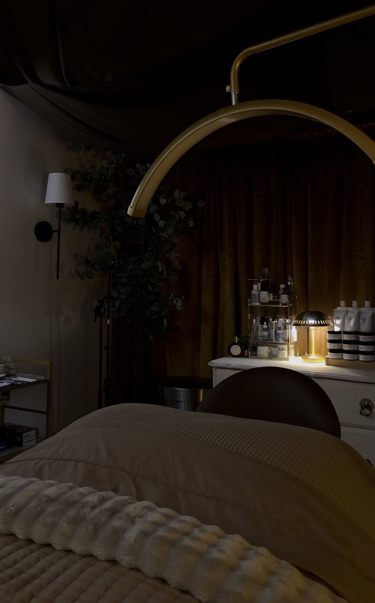 A dimly lit room with a bed, a side table, and a bar cart with bottles, against a curtain backdrop.