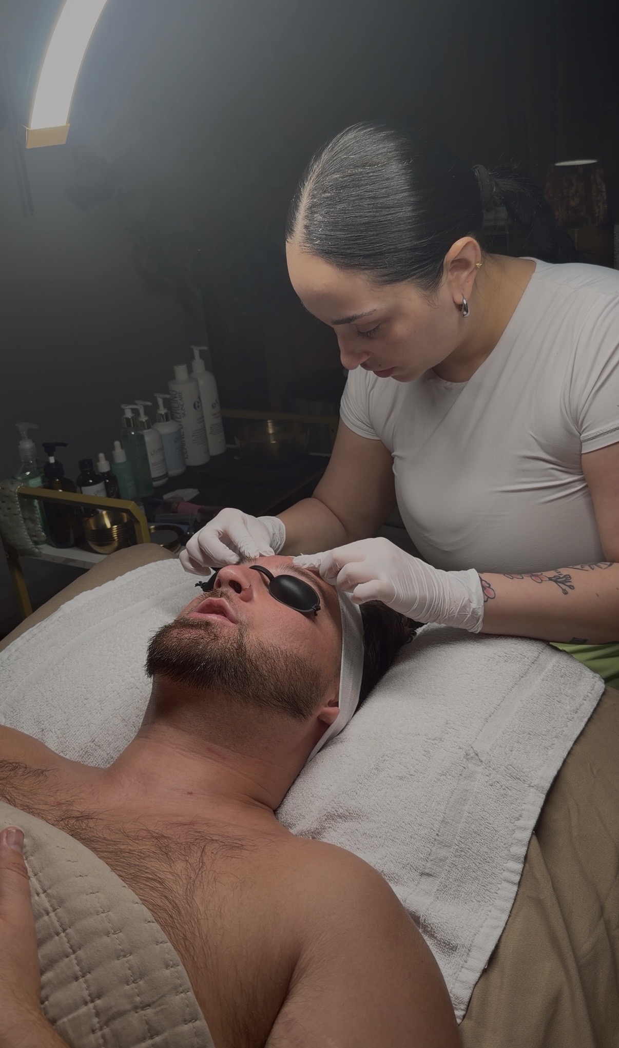 Woman with dark hair in a bun performs a facial treatment on a shirtless man lying on a bed, wearing eye protection.