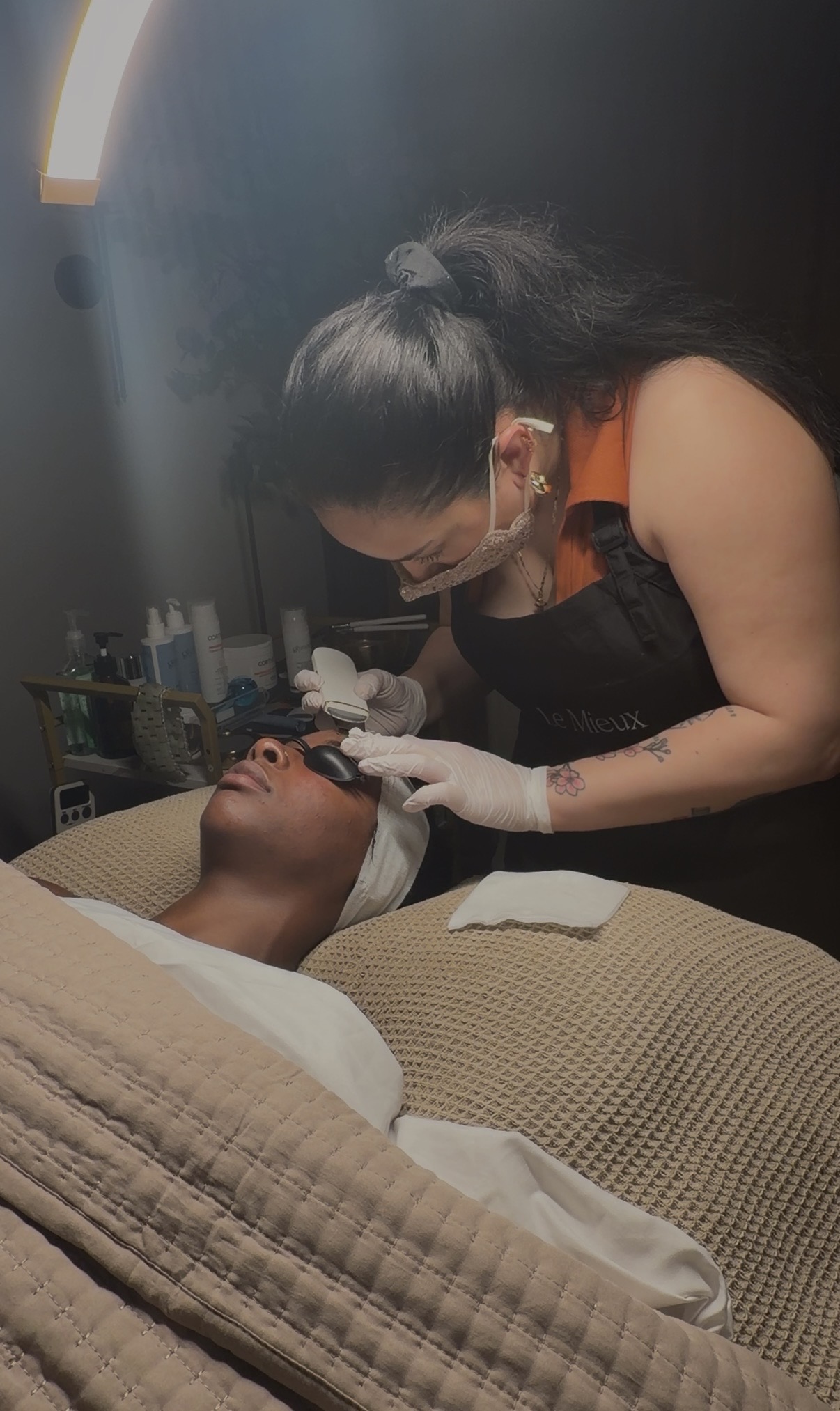 A woman wearing a mask and gloves performs a facial treatment on a person lying down with eyes closed, in a spa setting.
