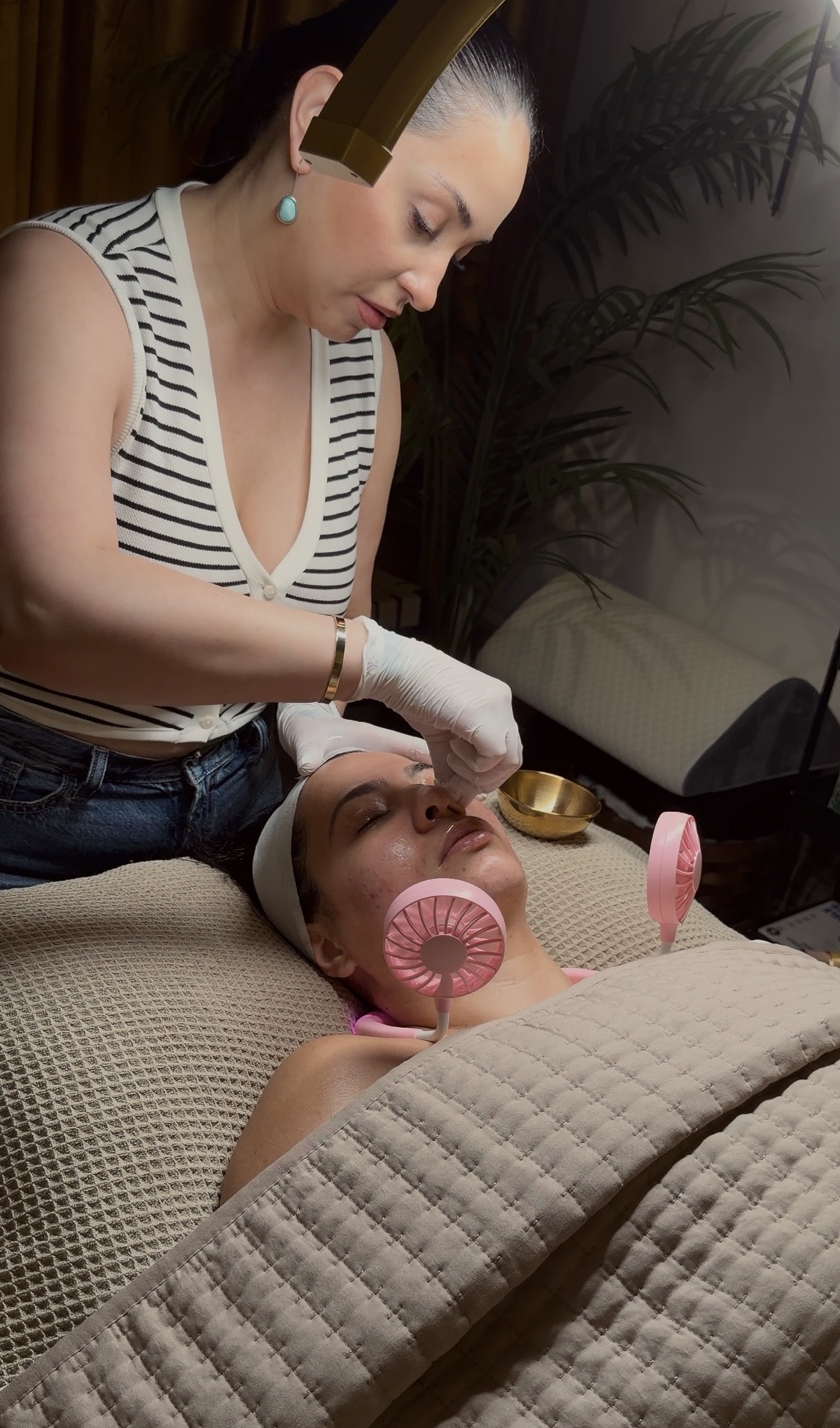 Woman providing facial treatment to a person lying on a bed, with pink facial mask and a pink fan, in a cozy room.
