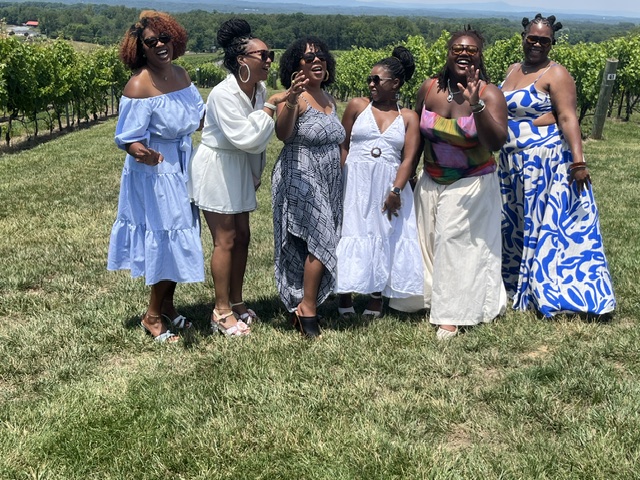 Group of seven women standing outdoors on grass with vineyard in background, smiling and talking, dressed in summer clothing.