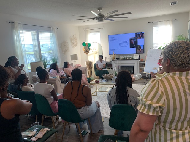 Group of people seated in a room watching a presentation on a large screen, with a person speaking at the front.