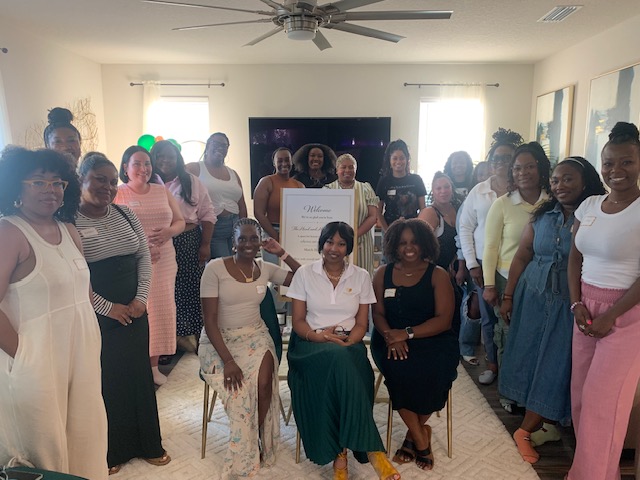 Group of women gathered in a living room, some sitting and some standing, with a large TV in the background.