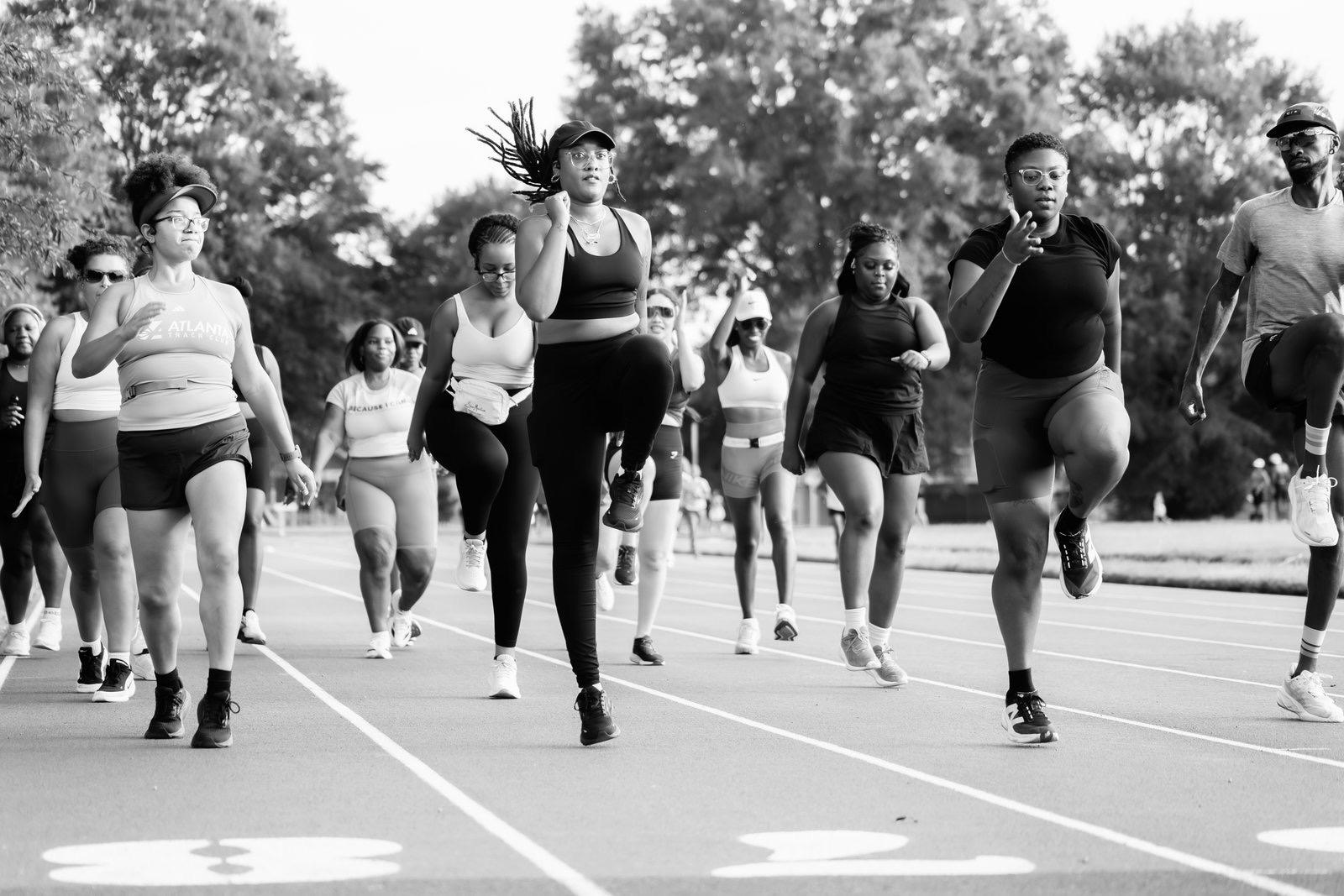 Group of women running outdoors on a track, trees in the background, black and white photo.