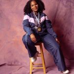 Young woman with curly hair sitting on a stool, wearing a navy and pink jacket, with a camera around her neck, smiling.