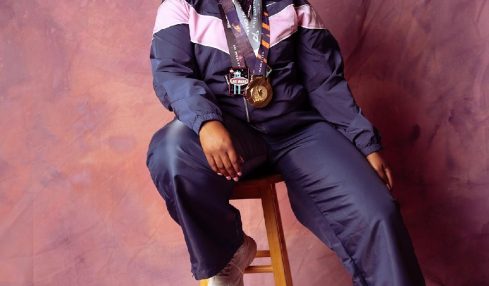 Young woman with curly hair sitting on a stool, wearing a navy and pink jacket, with a camera around her neck, smiling.
