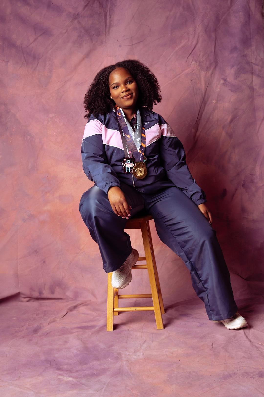 Young woman with curly hair sitting on a stool, wearing a navy and pink jacket, with a camera around her neck, smiling.