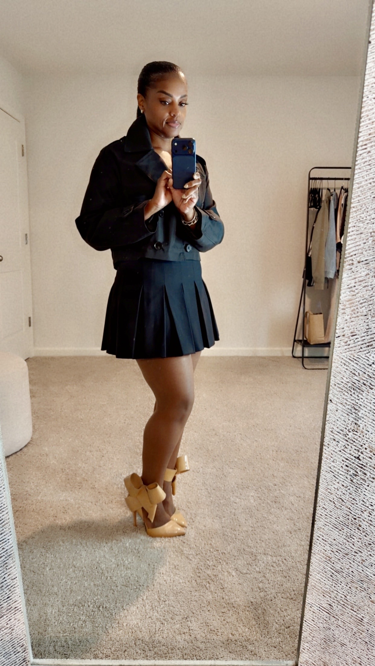 Person taking a mirror selfie in a room, wearing a dark jacket, pleated skirt, and high-heeled shoes, with a mask on.