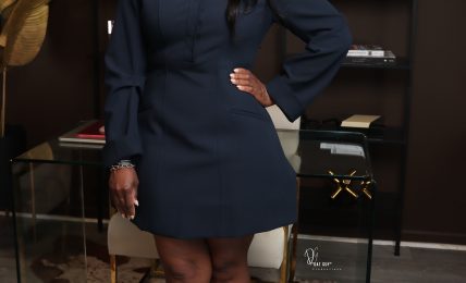 Woman in navy dress standing in a room with dark walls, shelves, and decorative items, smiling with one hand on her hip.