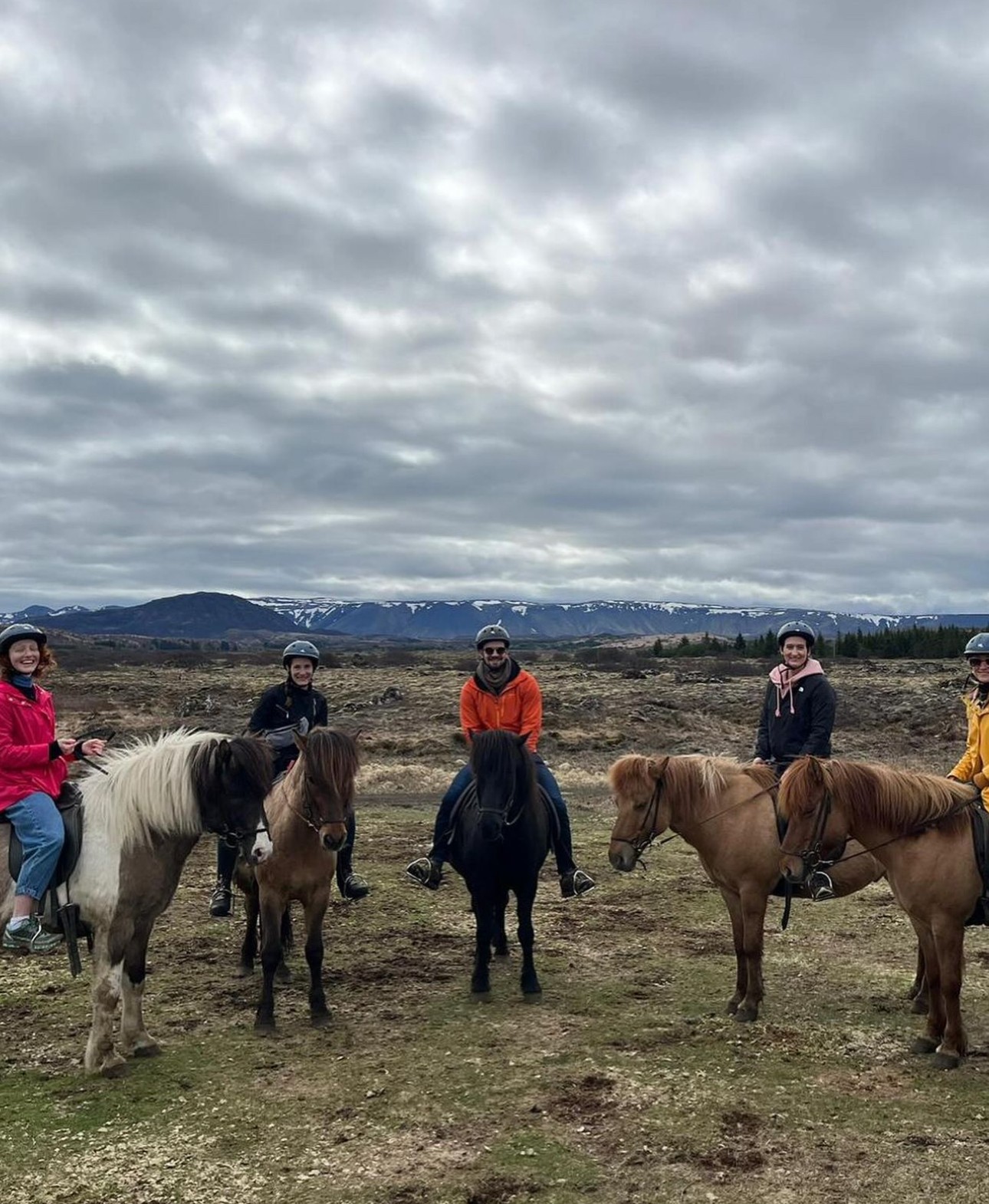Five people standing with horses outdoors under cloudy sky, landscape with distant hills in background.