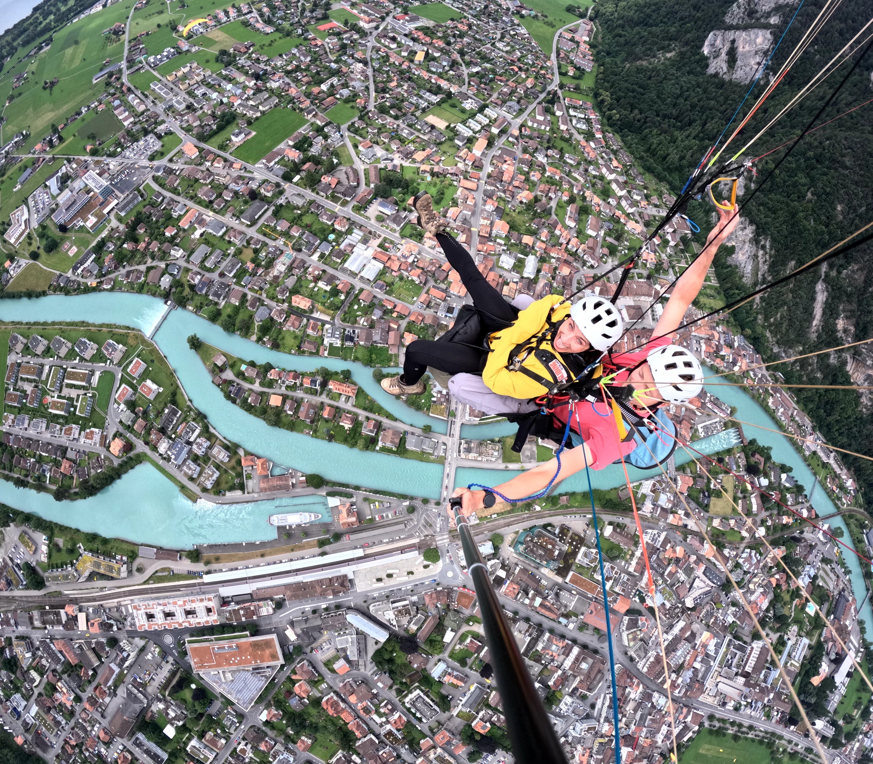 Two people are tandem paragliding over a city with rivers and green areas, wearing helmets and harnesses.