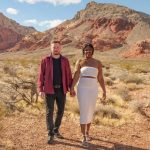 A couple holding hands walking in a desert landscape with mountains and blue sky in the background.