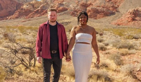 A couple holding hands walking in a desert landscape with mountains and blue sky in the background.
