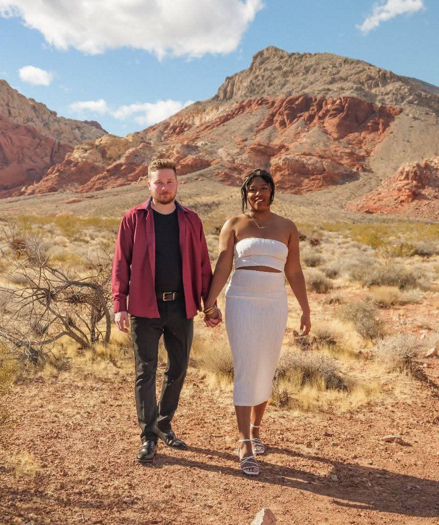 A couple holding hands walking in a desert landscape with mountains and blue sky in the background.