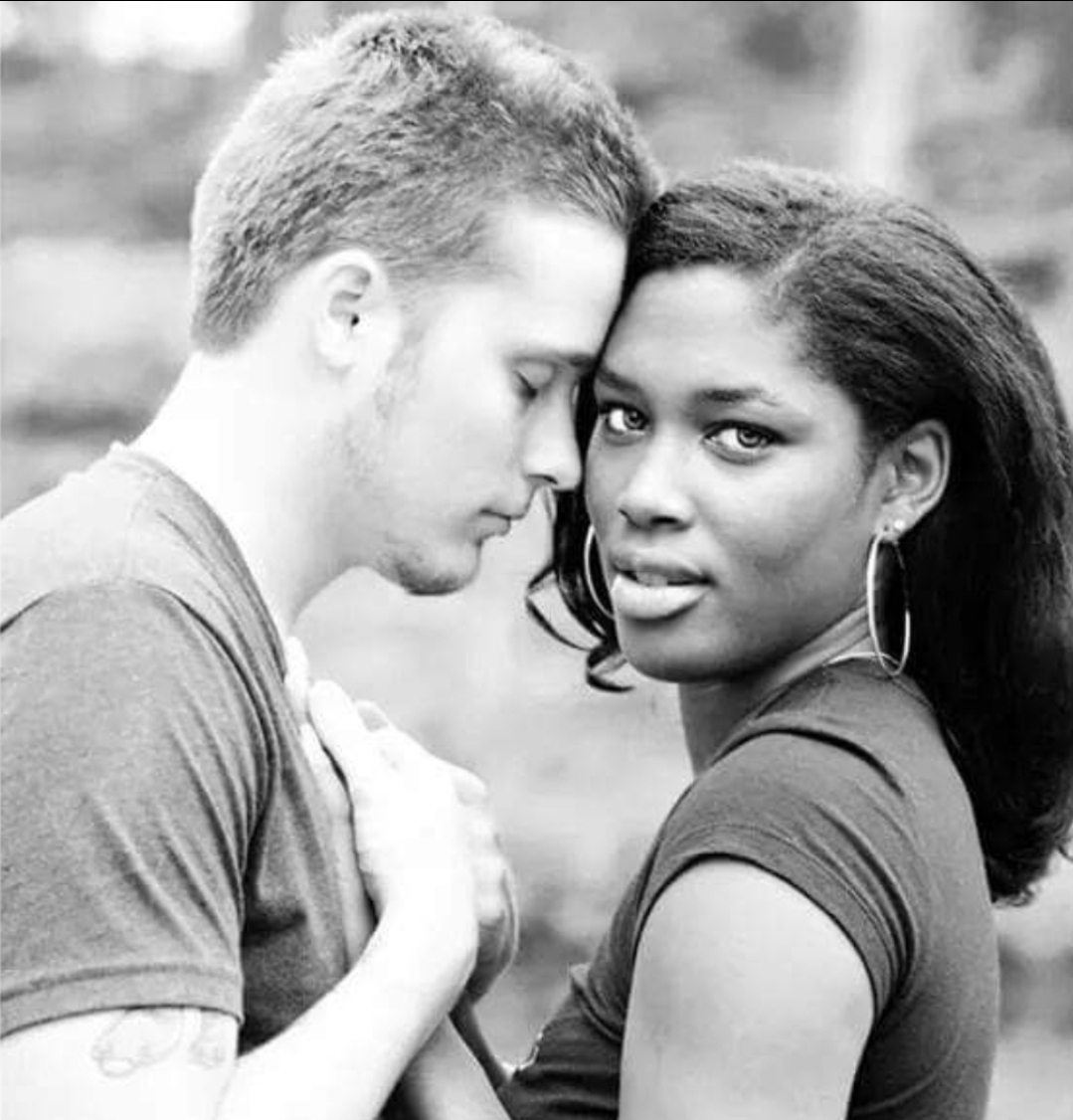 A man and woman face each other closely outdoors, the man with eyes closed and the woman looking at the camera.
