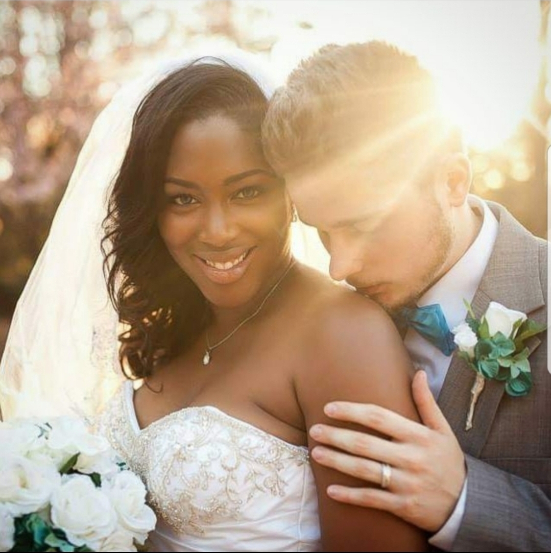 Bride and groom close together outdoors, smiling, with sunlight behind them, bride holding bouquet, groom wearing suit with boutonniere.