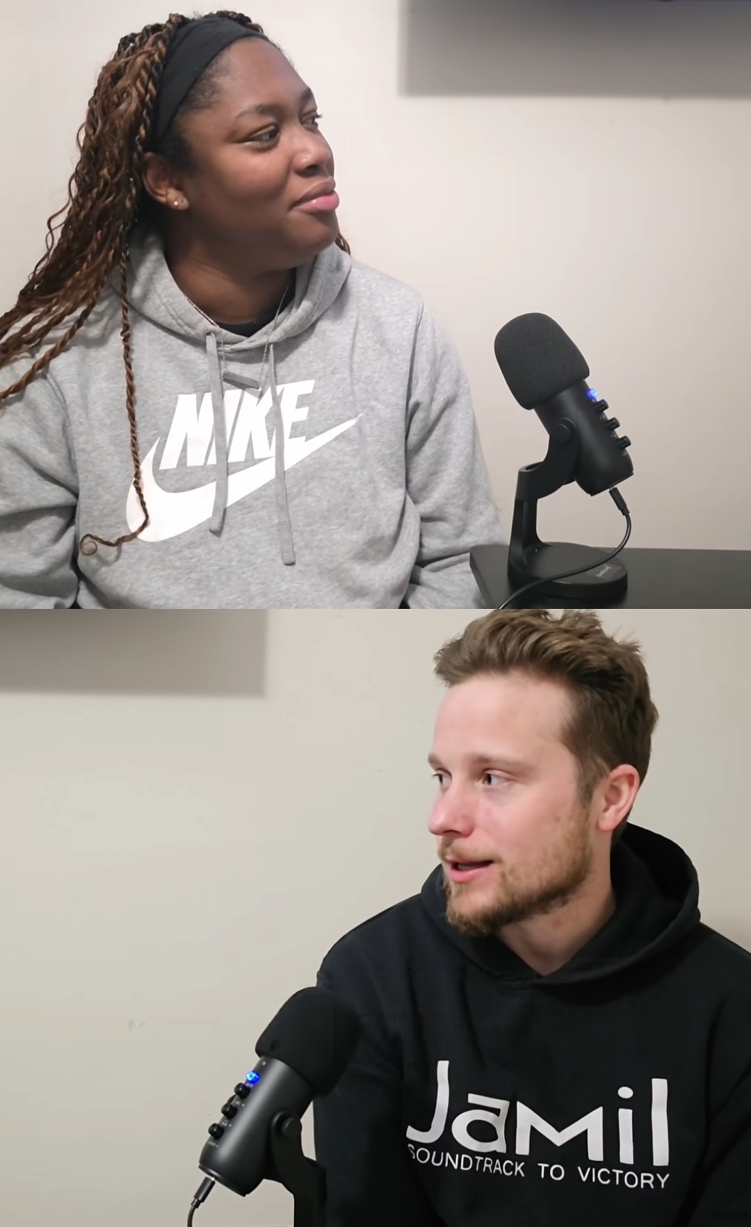 Two people sit at a table with microphones, engaged in conversation, against a plain background.