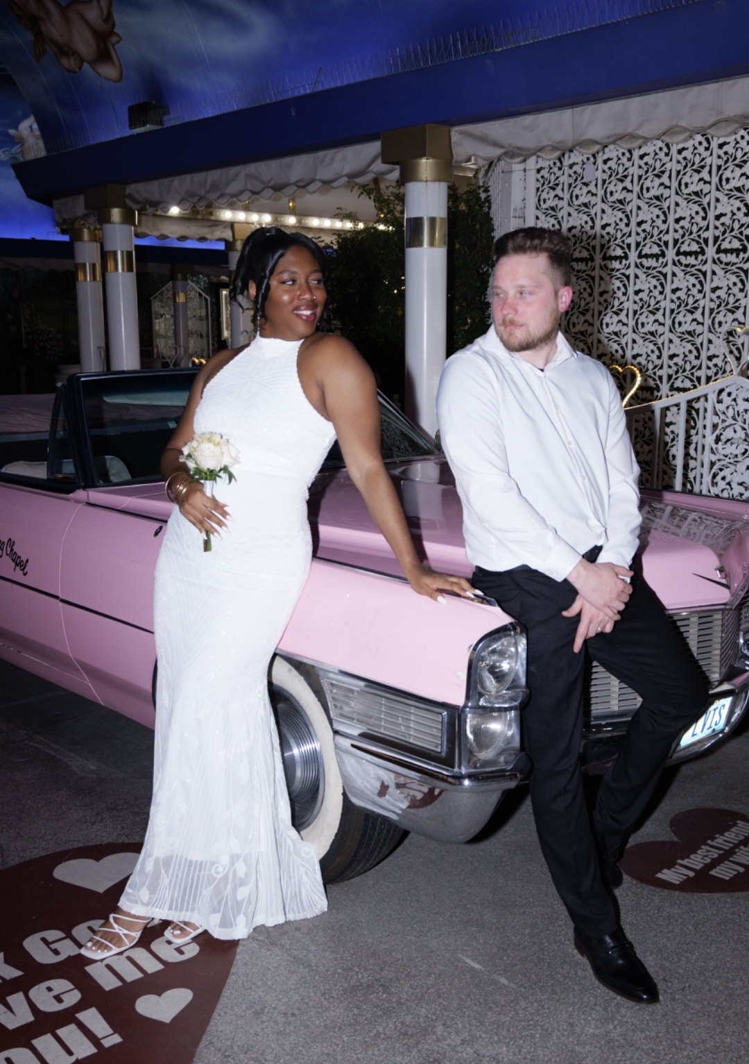 A woman in a white dress and a man in a white shirt and black pants pose in front of a pink vintage car at night. She is smiling and holding a bouquet, he is looking away. They are outdoors near a decorated structure.