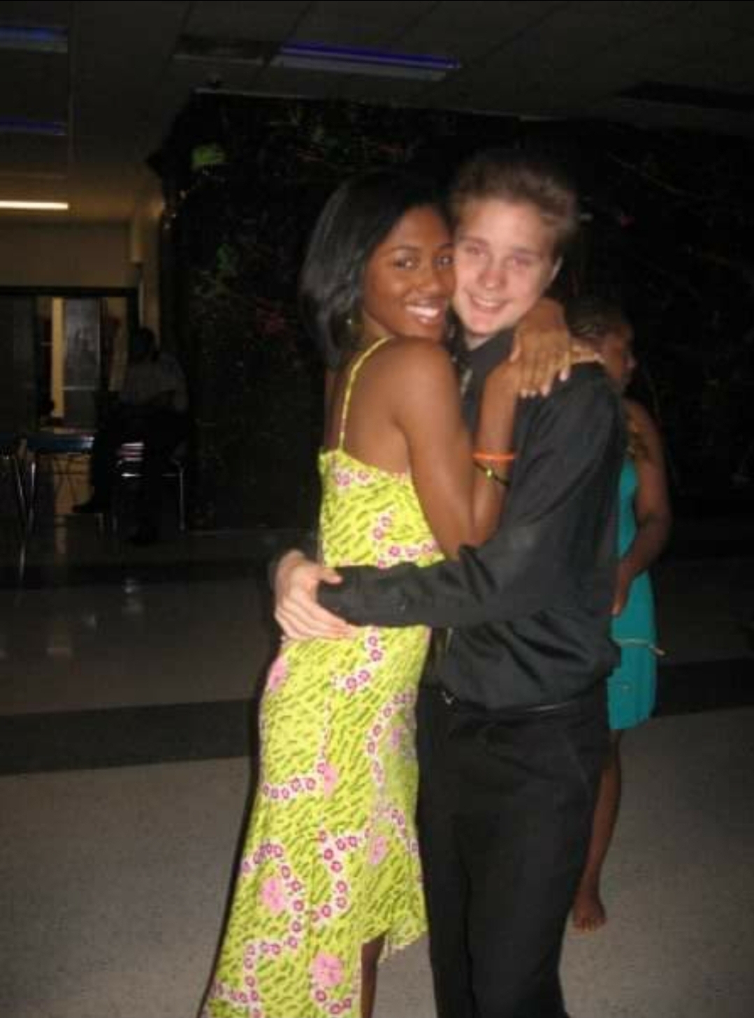 Two people hugging, smiling, in a dimly lit indoor setting, one wearing a bright yellow and pink dress, the other in dark clothing.