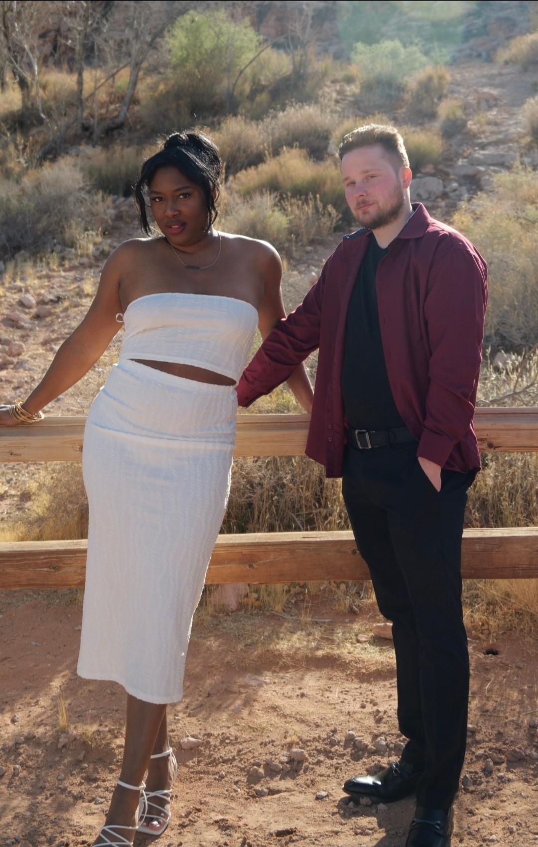 A woman and a man stand outdoors on a wooden platform with desert vegetation in the background. The woman wears a white strapless dress, and the man wears a maroon jacket and black pants. They are holding hands and looking at the camera.