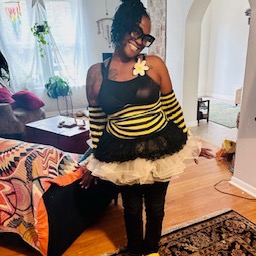 Person with glasses and a flower on face, wearing a black and yellow striped top with a ruffled skirt, standing indoors.