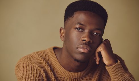 Young man with short hair wearing a brown sweater, resting his face on his hand, looking at the camera.
