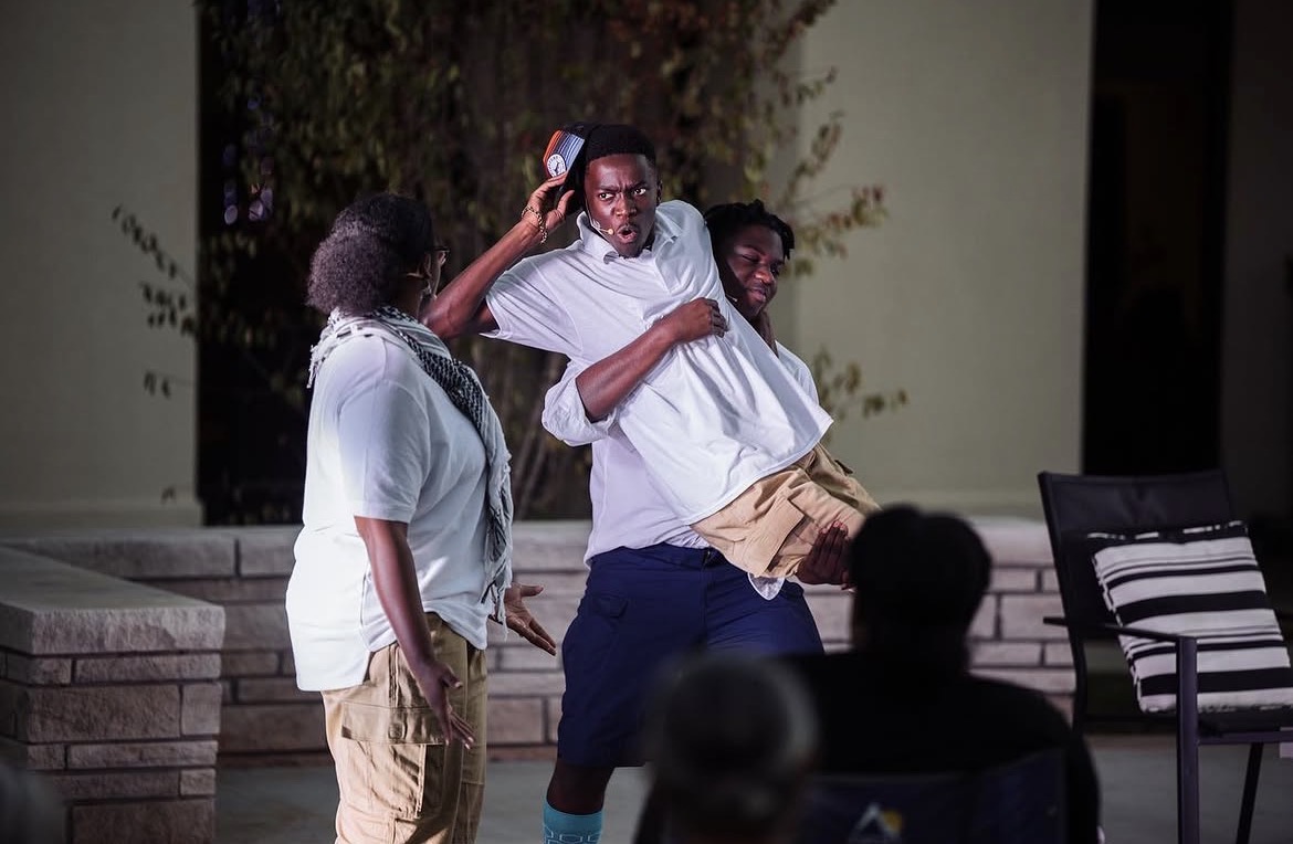 Three people outdoors at night, one holding a phone, others watching, with a chair nearby and a person in the foreground.
