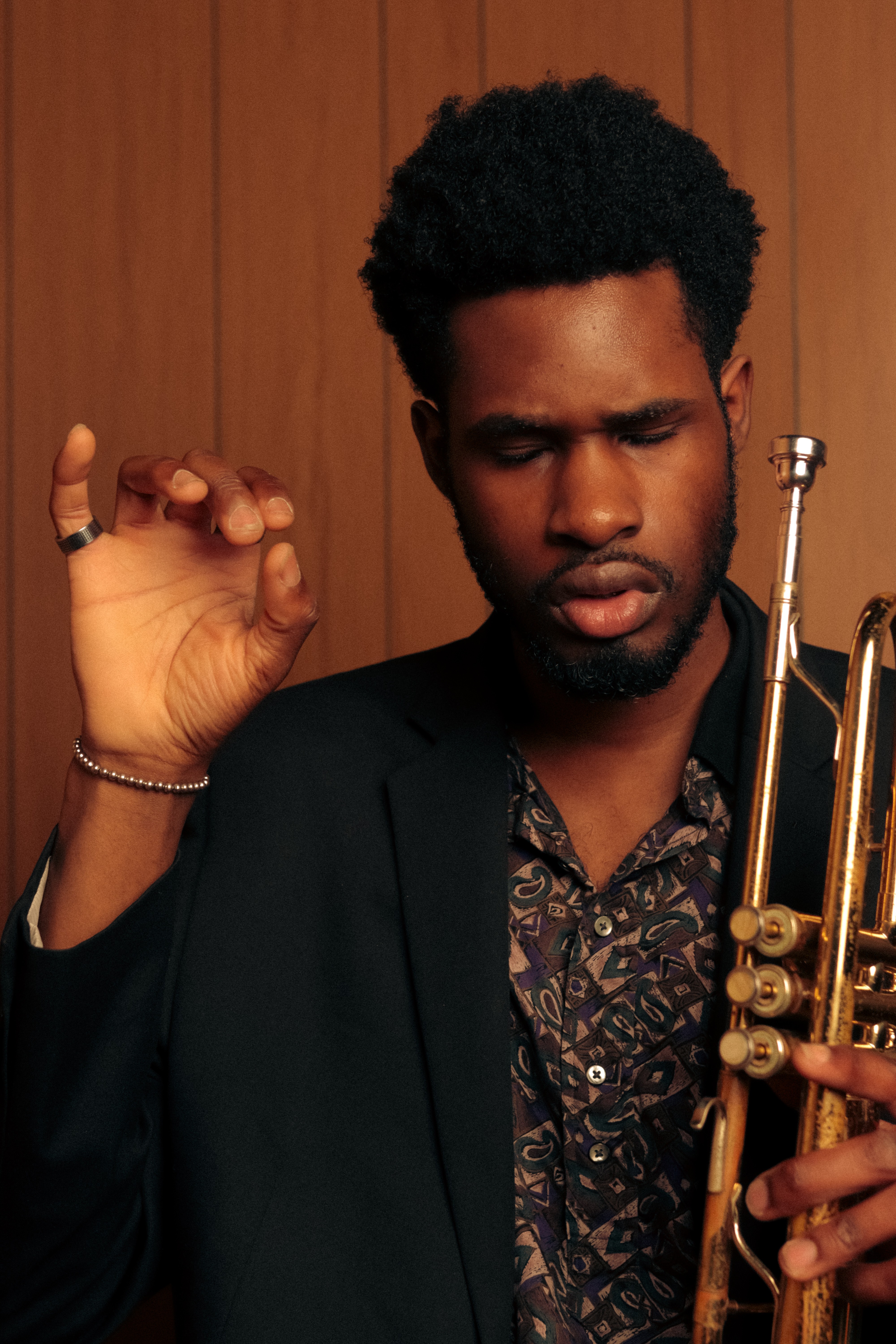 Man with curly hair and beard holding a trumpet, eyes closed, in front of wooden panel background.