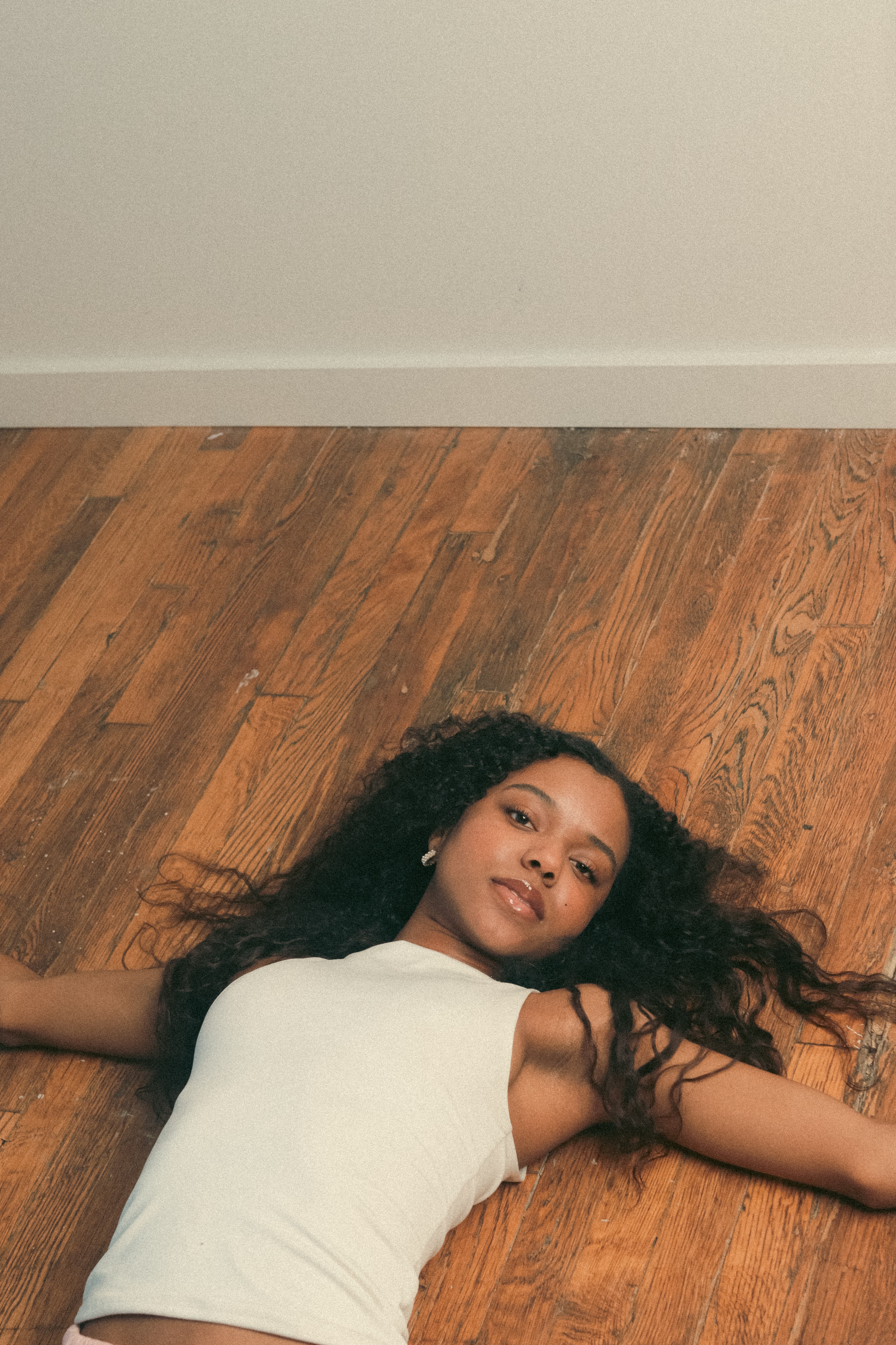 Young woman lying on wooden floor with arms outstretched, wearing a white sleeveless top, against a plain wall.